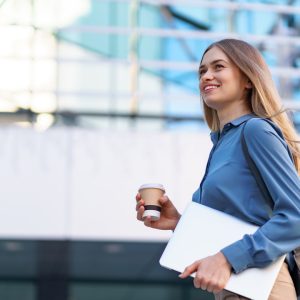 Blonde young woman smiling portrait holding laptop and coffee, wearing blue gentle shirt over modern building background